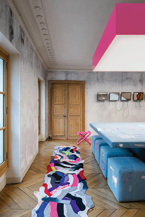 Installazione di Harry Nuriev con tappeto di calzini colorati cuciti sul parquet, divano modulare blu elettrico e soffitto con elemento rosa fluorescente in ambiente con pareti in intonaco grezzo
