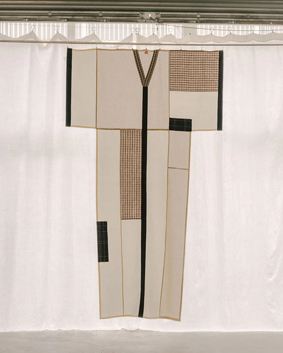 Tenda a forma di kimono oversize in lino naturale con patchwork di tessuti a quadretti neri e ocra appesa su fondale di lino bianco con monogrammi