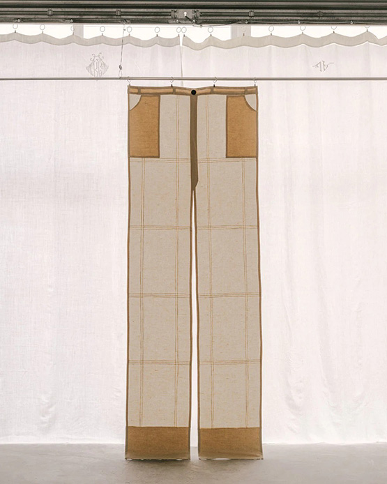 Tenda a forma di pantaloni a zampa in lino e iuta con quadretti ocra appesa con clip a barra metallica su fondale di lino bianco con monogrammi