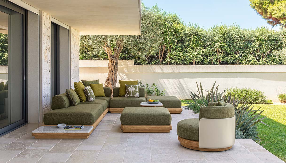 Sistema modulare Itaca Talenti in tessuto boucl&eacute; avorio con cuscini in tonalit&agrave; terracotta e rosa antico, tavolini rotondi in teak con piano in vetro, terrazza in pietra chiara con ulivo e siepe verde, porta scorrevole sul living sullo sfondo