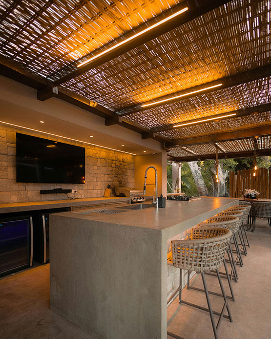 Angolo bar outdoor serale con bancone in cemento, sgabelli in rattan intrecciato, cantinetta a incasso e soffitto a boiserie illuminato a LED