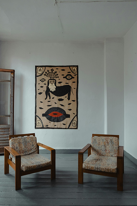Arazzo figurativo in beige, nero e rosso con motivi simbolici appeso su parete bianca tra due poltrone vintage in legno