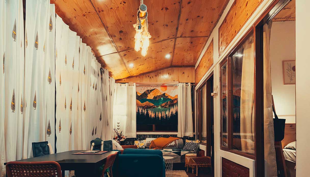 Arazzo con paesaggio montano dai colori intensi appeso come fulcro visivo in un soggiorno eclettico con soffitto in legno e tende bianche