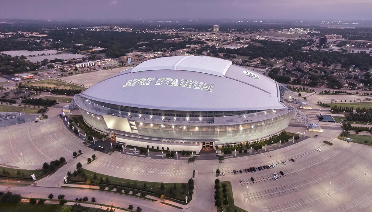 Vista aerea dell'AT&T Stadium di Arlington al crepuscolo, con il tetto retrattile chiuso e le luci del perimetro accese
