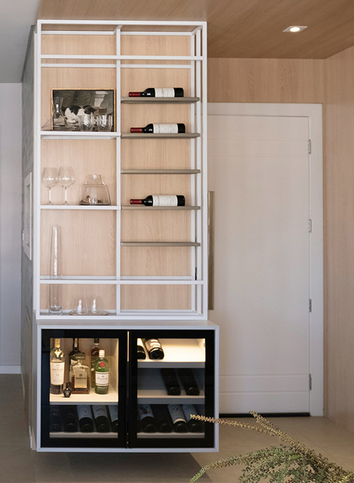 Angolo bar integrato in una libreria con struttura in metallo bianco, ripiani in legno chiaro per bottiglie e calici, e cantinetta refrigerata nella parte inferiore