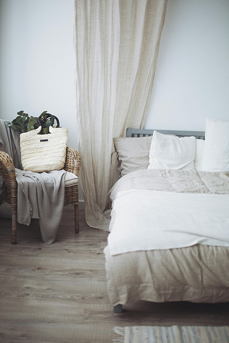 Camera da letto in toni neutri con letto in lino beige, poltrona in vimini e tenda in tessuto naturale che filtra la luce