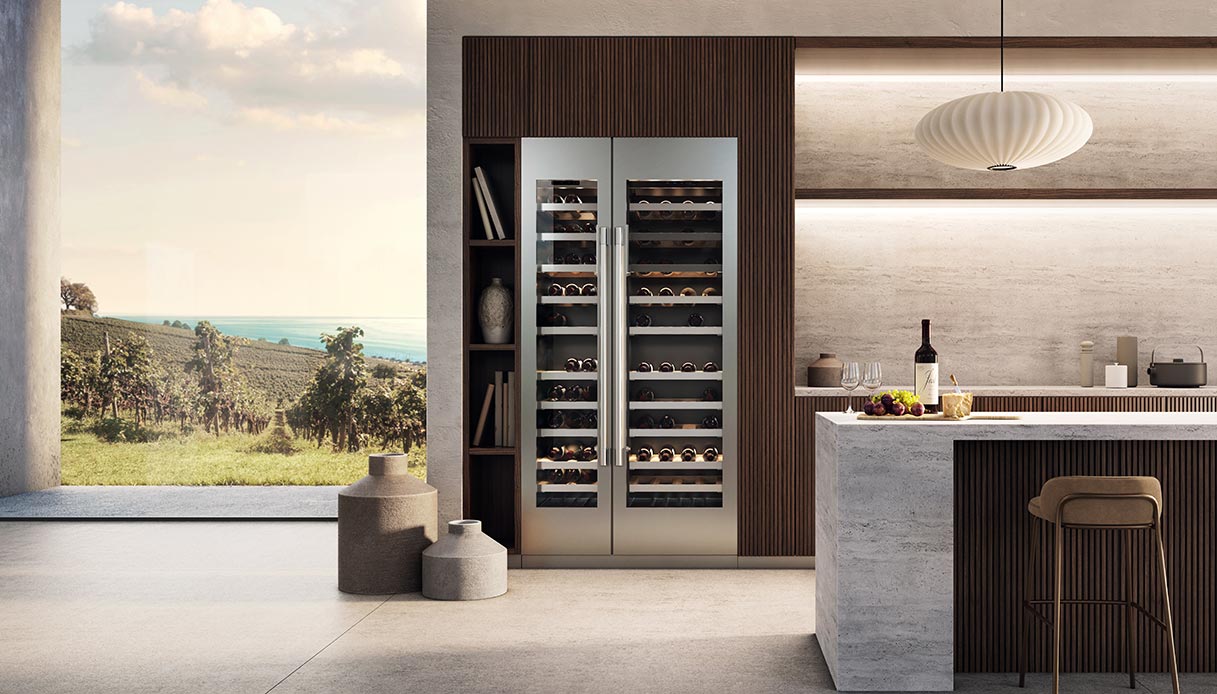 Cantinetta refrigerata a doppia colonna con ante in vetro e acciaio integrata in una parete in legno scuro scanalato, con isola in marmo e vista su vigneto