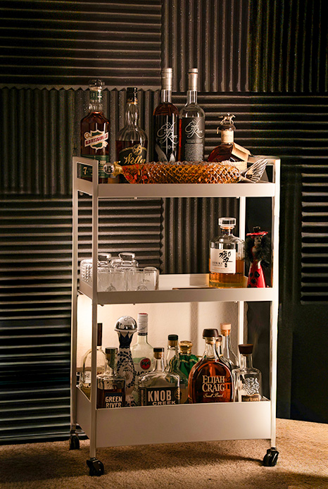 Carrello bar su ruote a tre ripiani in metallo con bottiglie di spirits, bicchieri e accessori da mixology su sfondo in lamiera ondulata