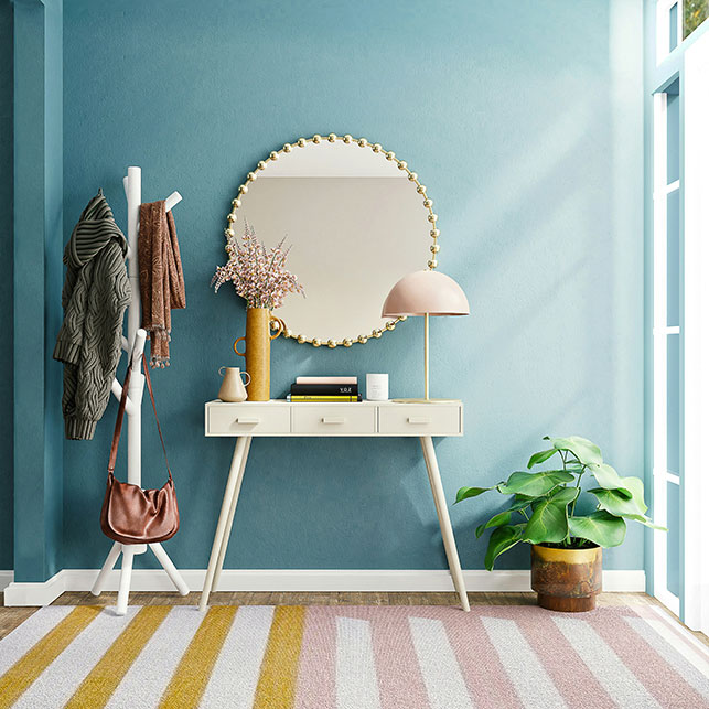 Ingresso con parete azzurra, consolle in legno chiaro, specchio rotondo con cornice dorata, lampada con paralume rosa cipria, appendiabiti bianco e tappeto a righe rosa giallo bianco