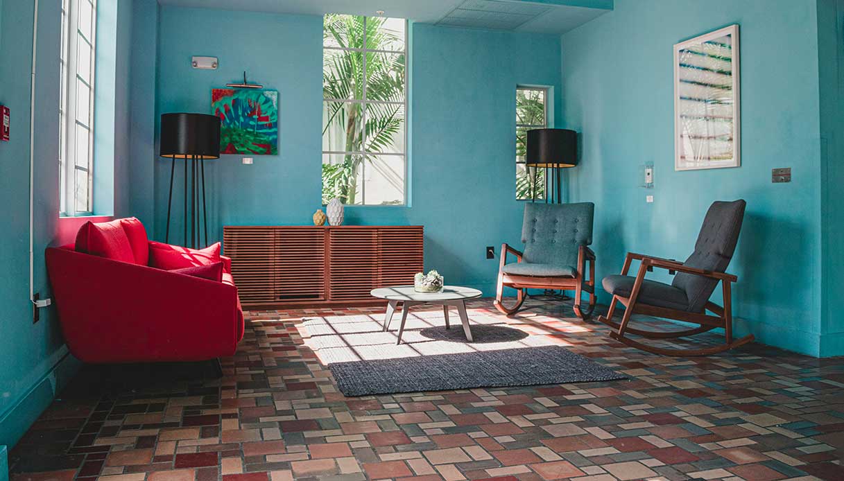 Soggiorno con pareti azzurro intenso, divano rosso brillante, sedie a dondolo grigio antracite, credenza in legno, pavimento in mattoni di cotto multicolore e piante tropicali