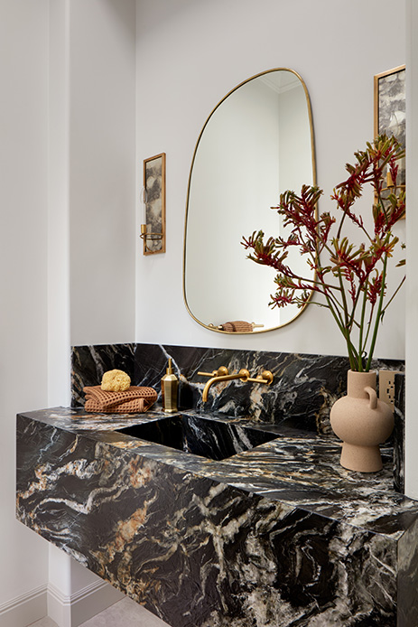 Bagno di servizio con lavabo monolitico in marmo nero con venature oro e grigio, rubinetteria a parete in ottone e specchio asimmetrico con cornice sottile dorata