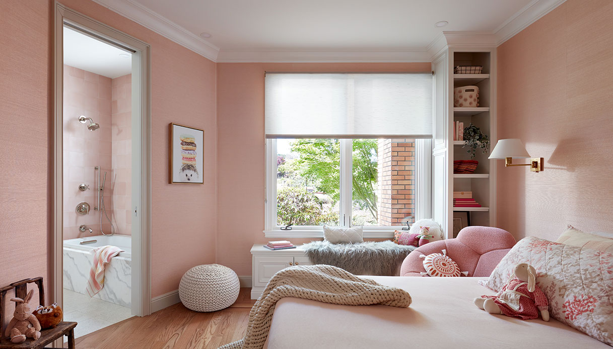 Camera da letto per bambini con pareti in carta da parati in sisal rosa, poltroncina boucl&eacute; rosa, panca sotto la finestra e bagno en suite con vasca rivestita in marmo bianco visibile sullo sfondo