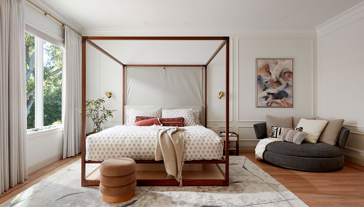Camera padronale con letto a baldacchino in legno scuro, applique in ottone sui montanti, divano rotondo grigio antracite, boiserie bianca a pannelli e quadro astratto con toni terracotta