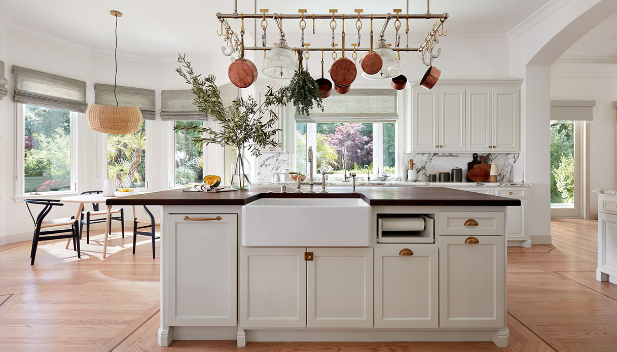 Cucina con ante verniciate in bianco crema, isola con piano in legno massiccio scuro, lavello farmhouse, struttura in ottone con pentole in rame appese e backsplash in marmo