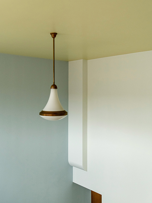 Angolo di stanza con soffitto in giallo ocra, lampada a sospensione in vetro opaco bianco con anello in ottone e pareti in azzurro chiaro
