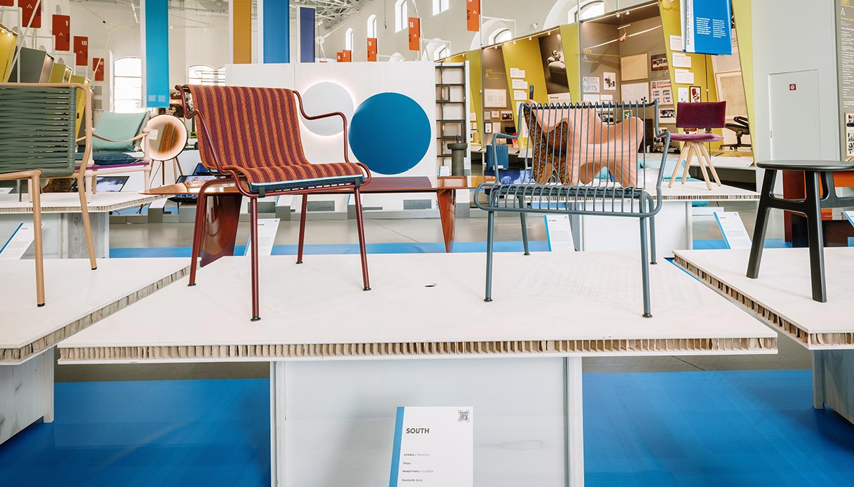 Allestimento mostra XXIX Compasso d'Oro con sedie di design su pedane bianche: in primo piano sedia rossa a righe e sedia grigio-azzurro a stecche, sullo sfondo altri prodotti premiati