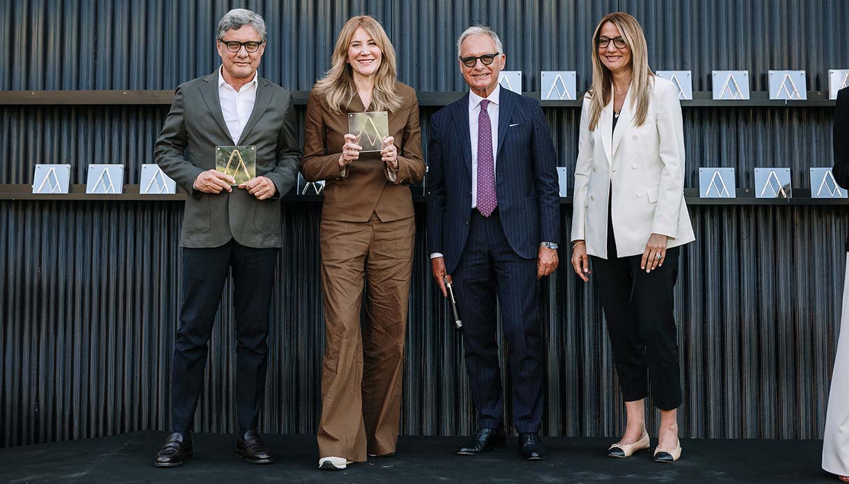 Quattro persone sul palco della cerimonia XXIX Compasso d'Oro ADI, due con targa dorata in mano, sullo sfondo parete metallica con targhe esposte