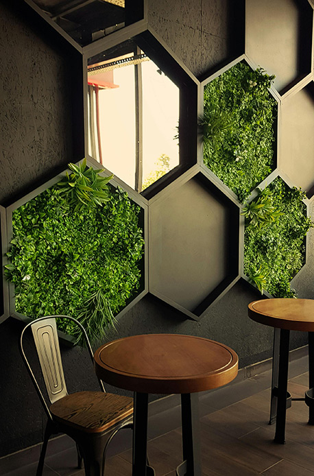 Giardino verticale con celle esagonali in metallo grigio su parete in cemento antracite, piante verdi vive e succulente in ogni cella, tavolino rotondo e sedia industriale in primo piano