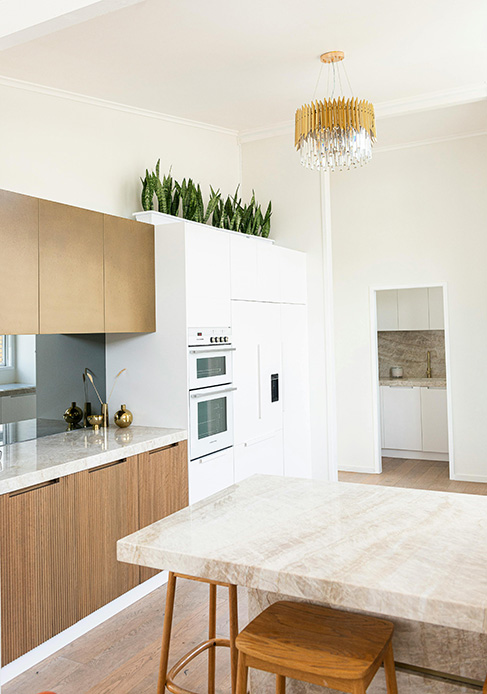 Cucina con pensili in legno bronzo e bianchi, sansevierie rigogliose sopra i pensili, isola con piano in marmo beige, lampadario in ottone e cristallo e parquet chiaro