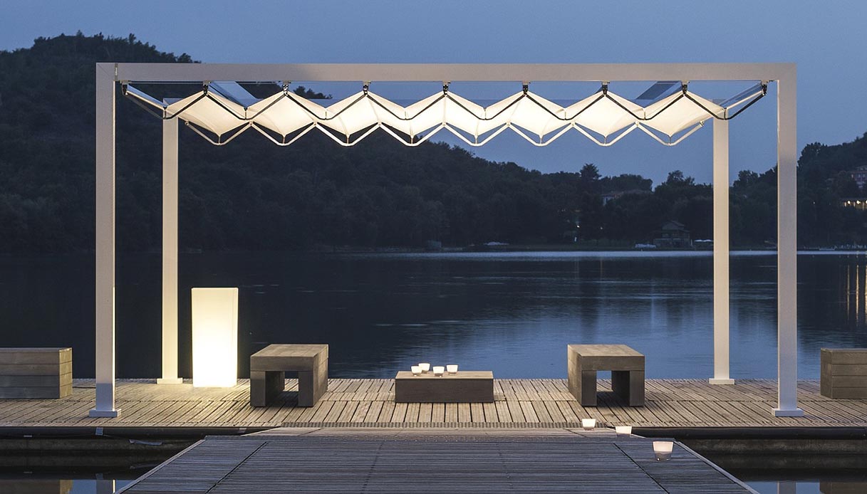 Pergola Gibus Med Open Fly con telo scorrevole bianco illuminato e struttura in alluminio bianco, su pontile in legno con vista su lago e colline al crepuscolo