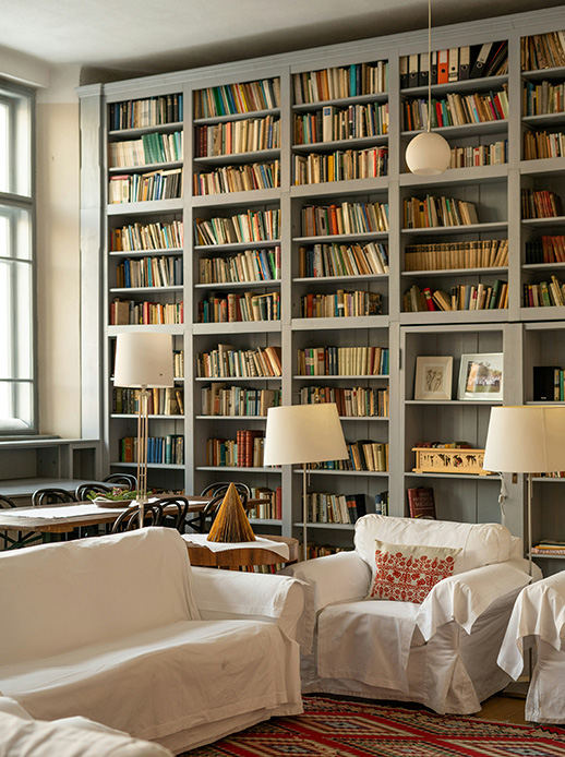 Soggiorno con libreria a parete intera grigio tortora piena di libri, piantane con paralume bianco, divano e poltrona bianchi con cuscino ricamato rosso e tappeto folk