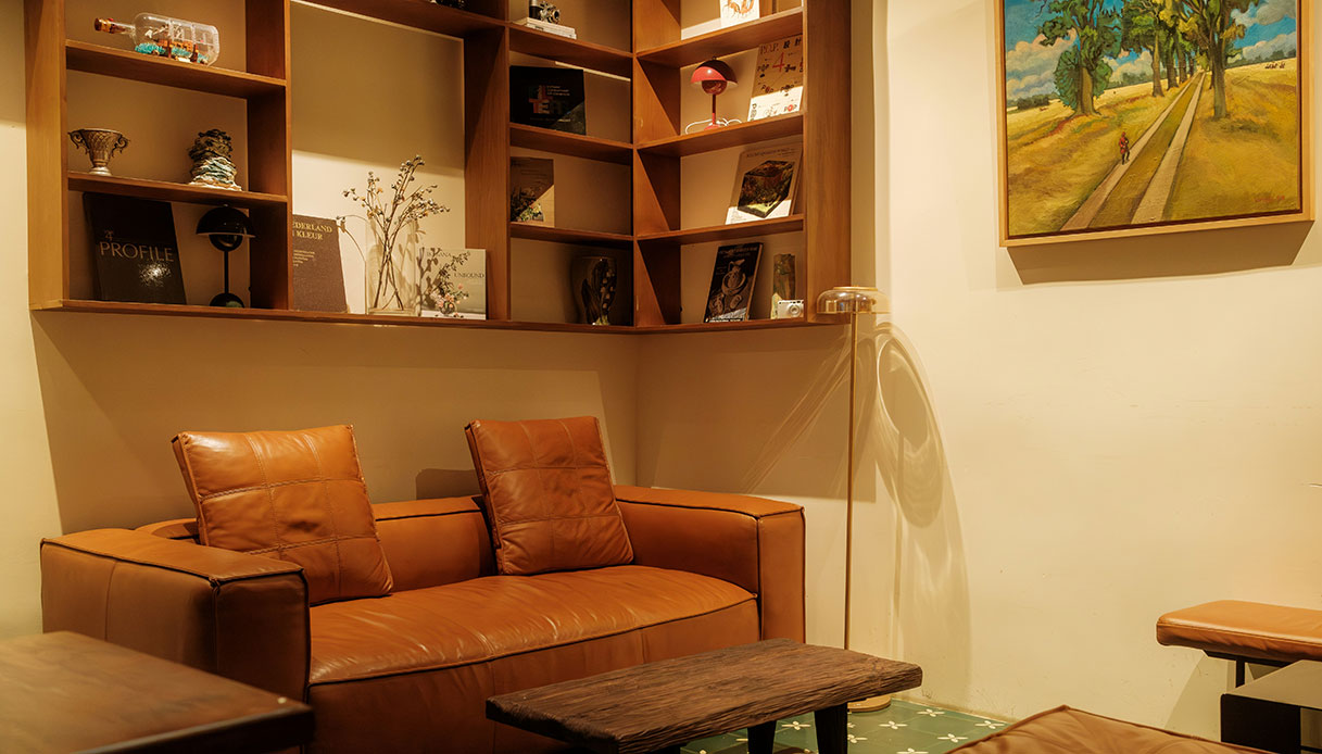 Soggiorno con libreria a mensole in legno con libri alternati a oggetti decorativi e piante secche, divano in pelle cognac e tavolino in legno scuro