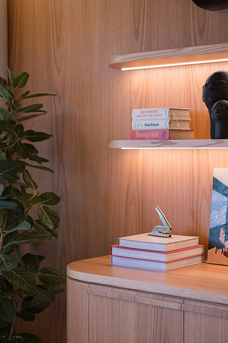 Dettaglio di mensole in legno chiaro con bordi arrotondati, retroilluminate a LED, con libri e oggetti decorativi su parete in legno