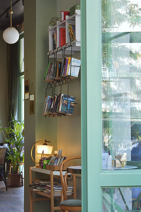 Porta-libri a tre ripiani in metallo nero sospeso sopra uno stipite di porta su parete verde, con libri e riviste a vista, lampada sferica e piante sullo sfondo