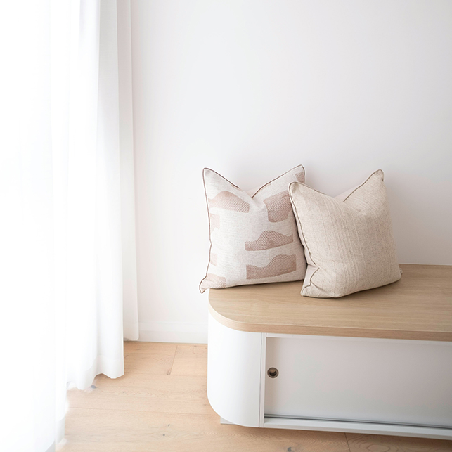 Panca con piano in legno chiaro e contenitore inferiore in bianco con sportello, con due cuscini in lino beige e rosa, in angolo luminoso con tende bianche