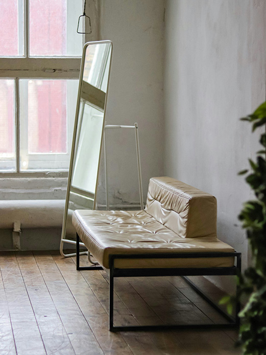 Panca trapuntata in ecopelle beige con struttura in metallo nero, affiancata da uno specchio a figura intera su cavalletto bianco, in ambiente con pareti in stucco grezzo