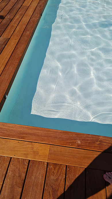 Dettaglio angolo piscina con decking in legno color miele caldo, acqua chiara con riflessi bianchi e bordo in resina azzurra