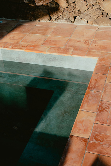 Bordo piscina con piastrelle di terracotta ruggine, acqua scura con riflessi e muro di pietra lavica grezza sullo sfondo