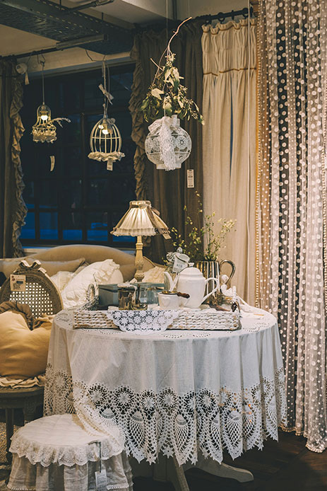Allestimento vintage con tavolo rotondo coperto da tovaglia in pizzo sangallo, tende a pois, gabbiette decorative appese e lampada da tavolo ottone