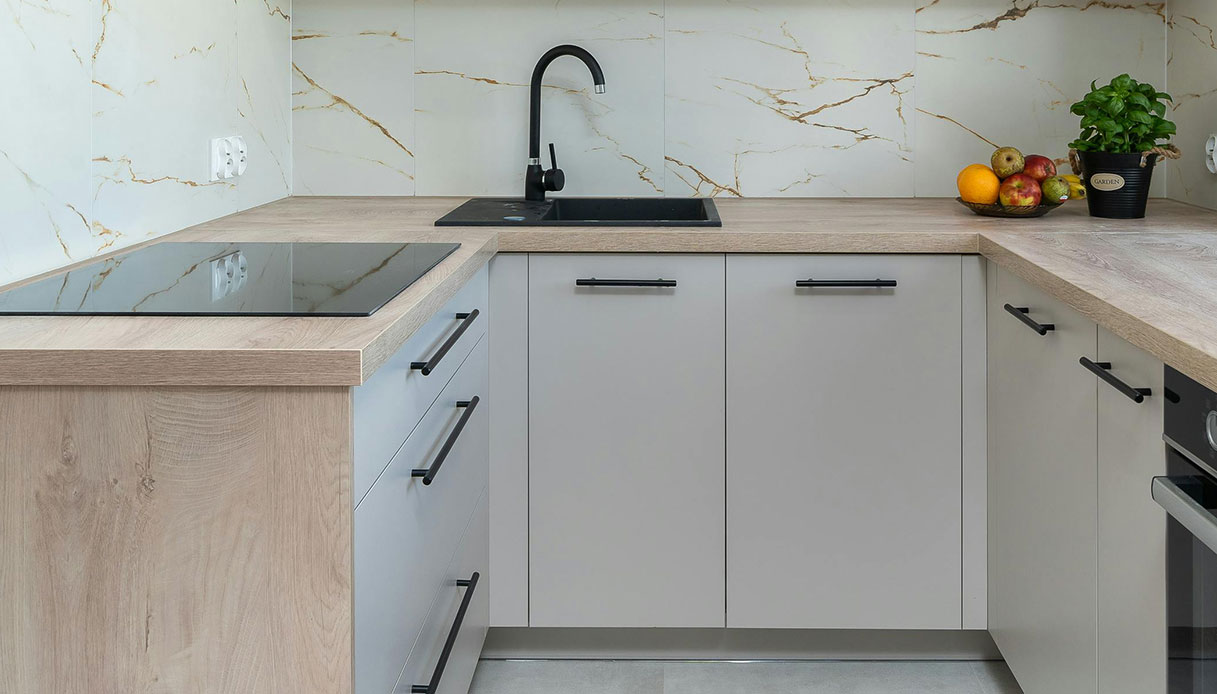 Cucina ad angolo con ante lisce grigio tortora, maniglie a barra in nero opaco, piano in legno rovere e paraschizzi in ceramica effetto marmo bianco con venature oro