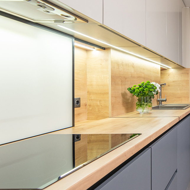 Cucina con paraschizzi in vetro extra-chiaro con pannello in legno naturale, striscia LED sottopensile, piano in legno chiaro e ante grigio chiaro