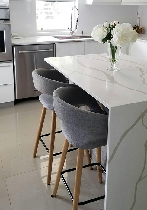 Isola cucina con piano in quarzo bianco con venature grigie, sgabelli imbottiti grigi con gambe in legno chiaro e vaso con peonie bianche