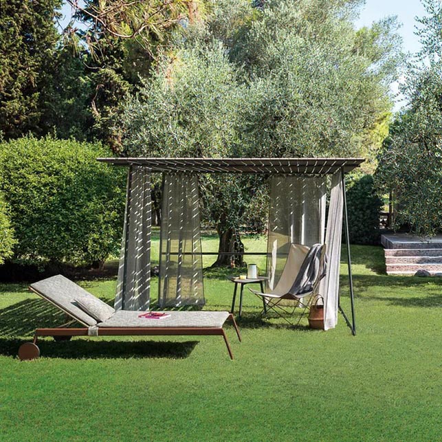 Gazebo Ombrina di Roda in tubolare di acciaio verniciato con tende fluttuanti in tessuto grigio, su prato verde con ulivi e siepi sullo sfondo
