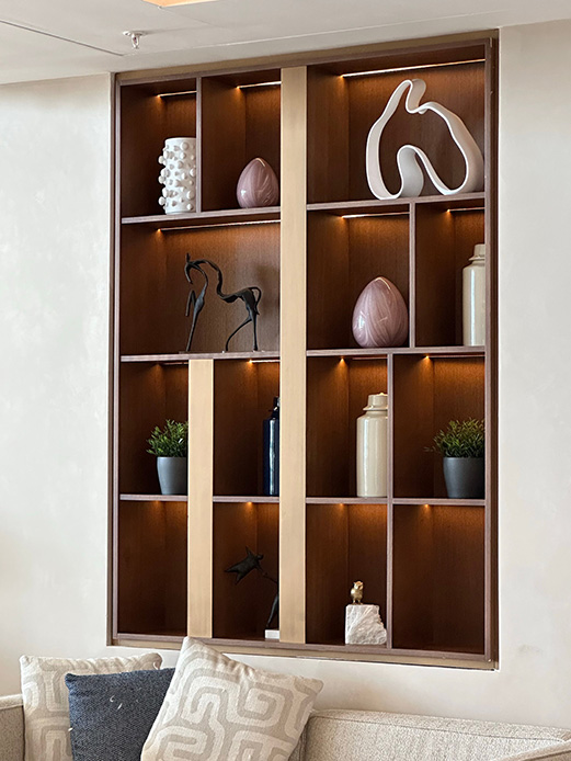 Sistema a muro in legno scuro con retroilluminazione calda, vani di dimensioni differenti esposti con sculture bianche, vasi rosa e piante, sopra un divano