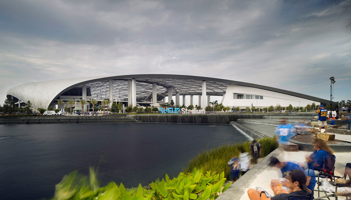 Vista esterna del SoFi Stadium di Inglewood con la copertura semitrasparente sorretta da colonne, uno specchio d'acqua in primo piano e tifosi lungo la riva