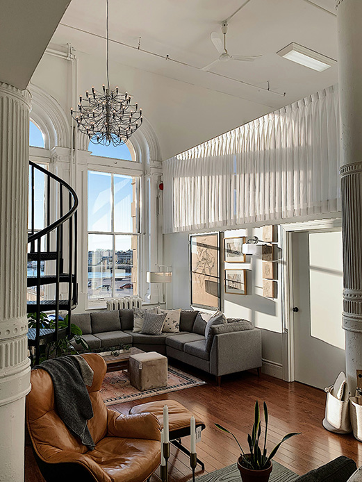 Loft con soffitto alto, tende plissettate bianche su finestre ad arco, scala elicoidale in ferro, divano grigio e poltrona in pelle marrone