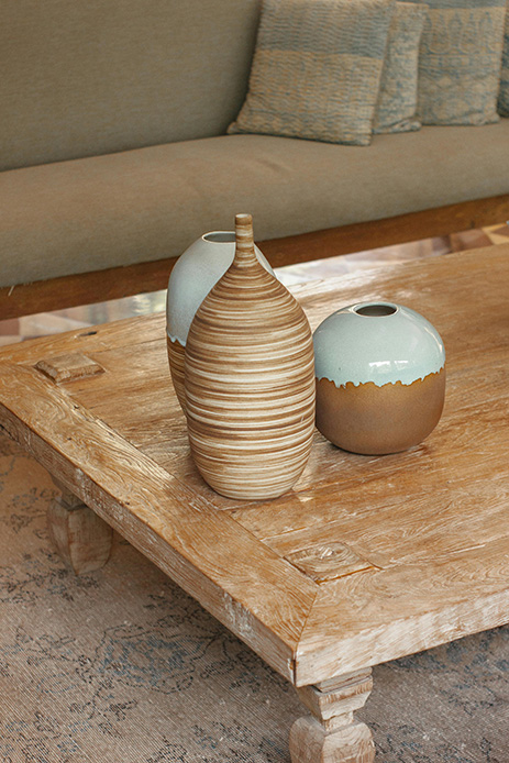 Due vasi in ceramica con smalto bicolore sabbia e azzurro su tavolino in legno grezzo consumato, con divano in tessuto neutro sullo sfondo