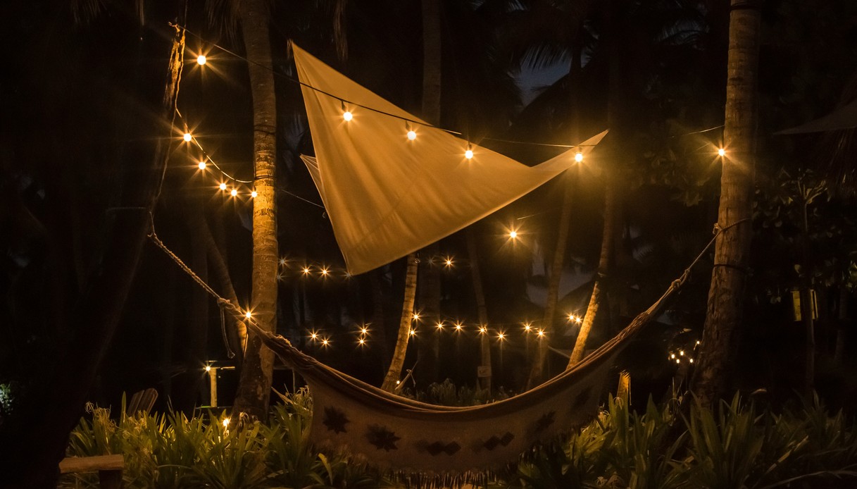 Amaca vuota tra le palme di notte, illuminata da una fila di lucine e una tenda a vela triangolare