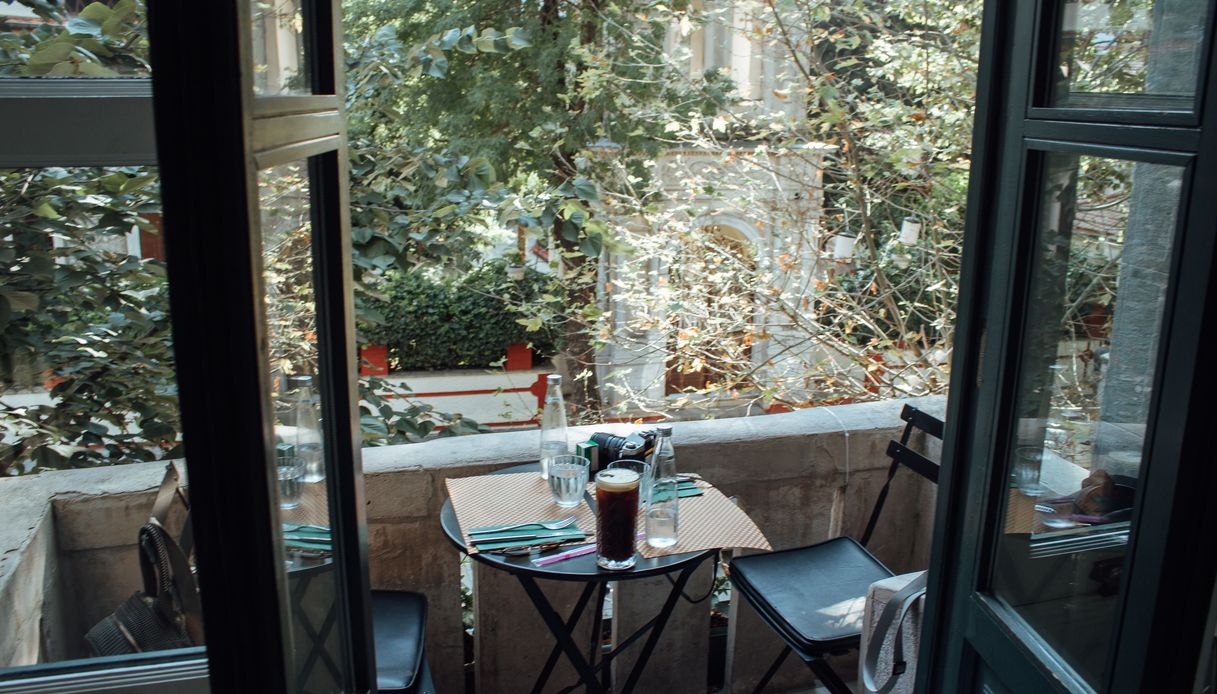 Piccolo balcone con tavolino tondo apparecchiato, sedie pieghevoli nere e vista su alberi e un edificio storico