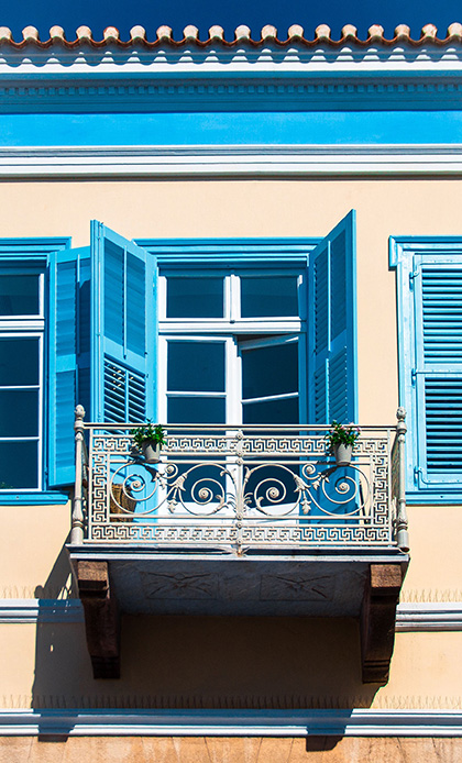 Balcone in ferro battuto lavorato con motivi decorativi, persiane blu aperte su facciata color sabbia