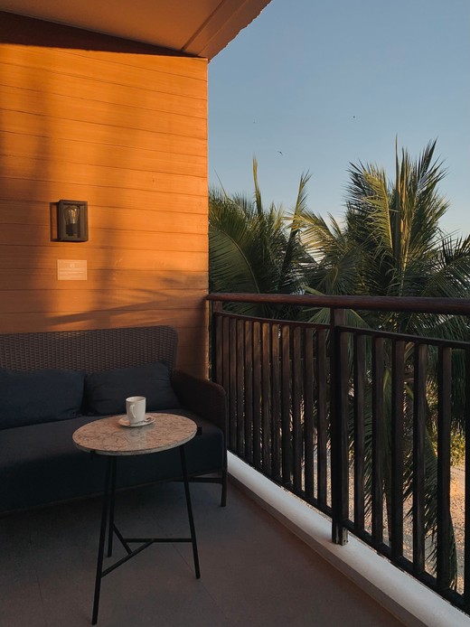 Balcone al tramonto con divano in vimini scuro, tavolino tondo con piano in pietra e tazza, ringhiera in legno e palme sullo sfondo
