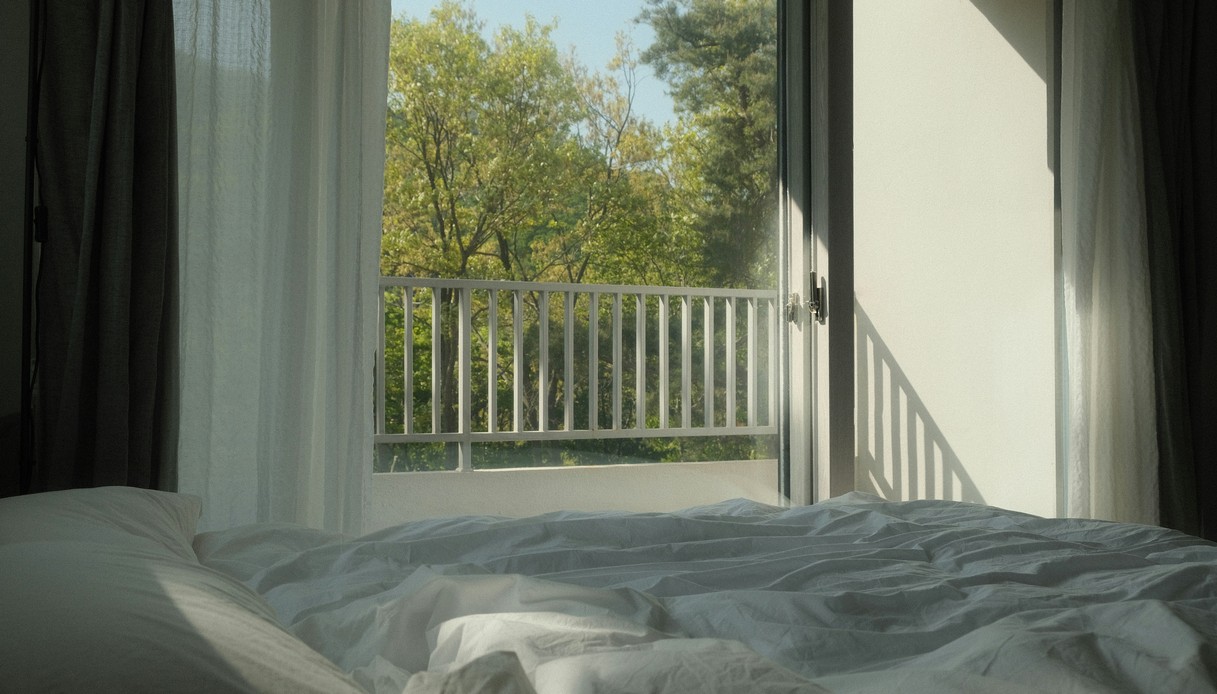 Vista dal letto verso una porta finestra aperta su un balcone con ringhiera bianca e alberi verdi, tende leggere ai lati