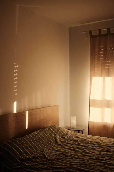 Camera da letto al tramonto con testata in legno, luce calda che filtra da tende doppie e comodino con lampada