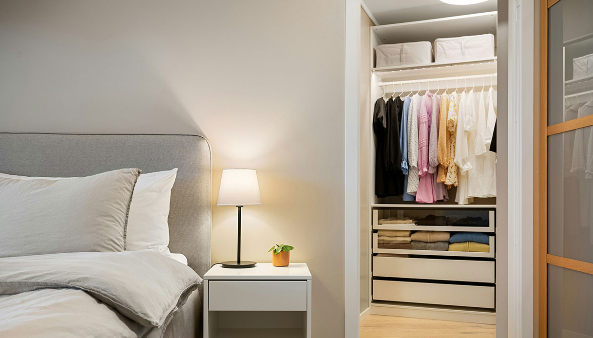 Comodino con lampada accesa accanto al letto, con armadio a vista contenente vestiti colorati appesi e cassetti aperti