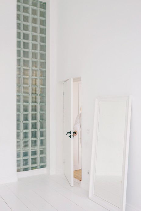 Corridoio bianco con parete in vetrocemento che filtra la luce, specchio a figura intera e porta socchiusa