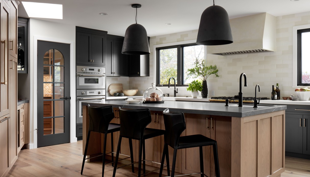 Cucina con isola in rovere chiaro, piano scuro, tre sgabelli neri e due lampade a sospensione nere, ante scure sullo sfondo